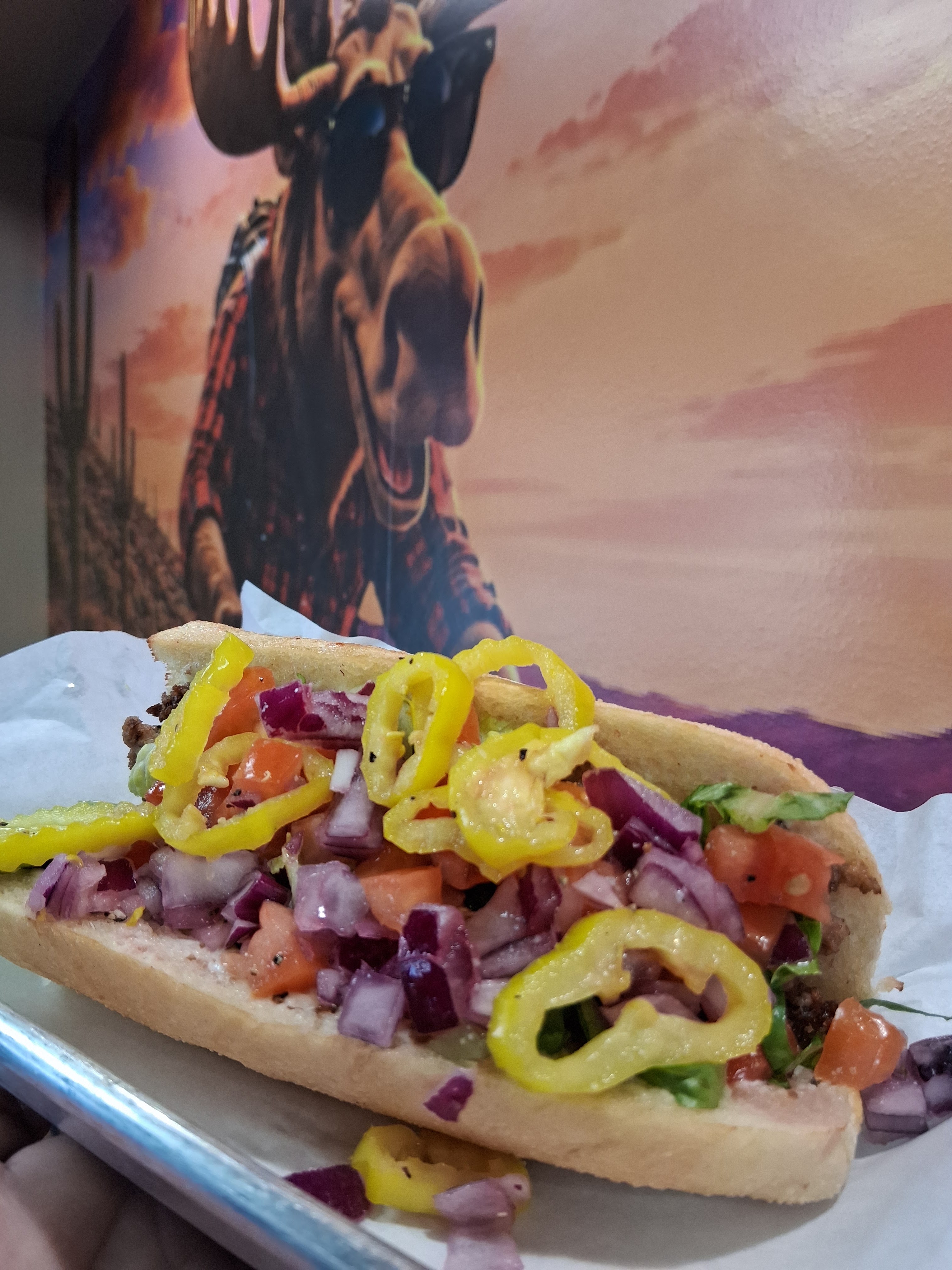 A hot dog topped with chopped tomatoes, onions, lettuce, and yellow pepper rings, served on a paper-lined tray, with a colorful mural of a moose wearing sunglasses in the background.
