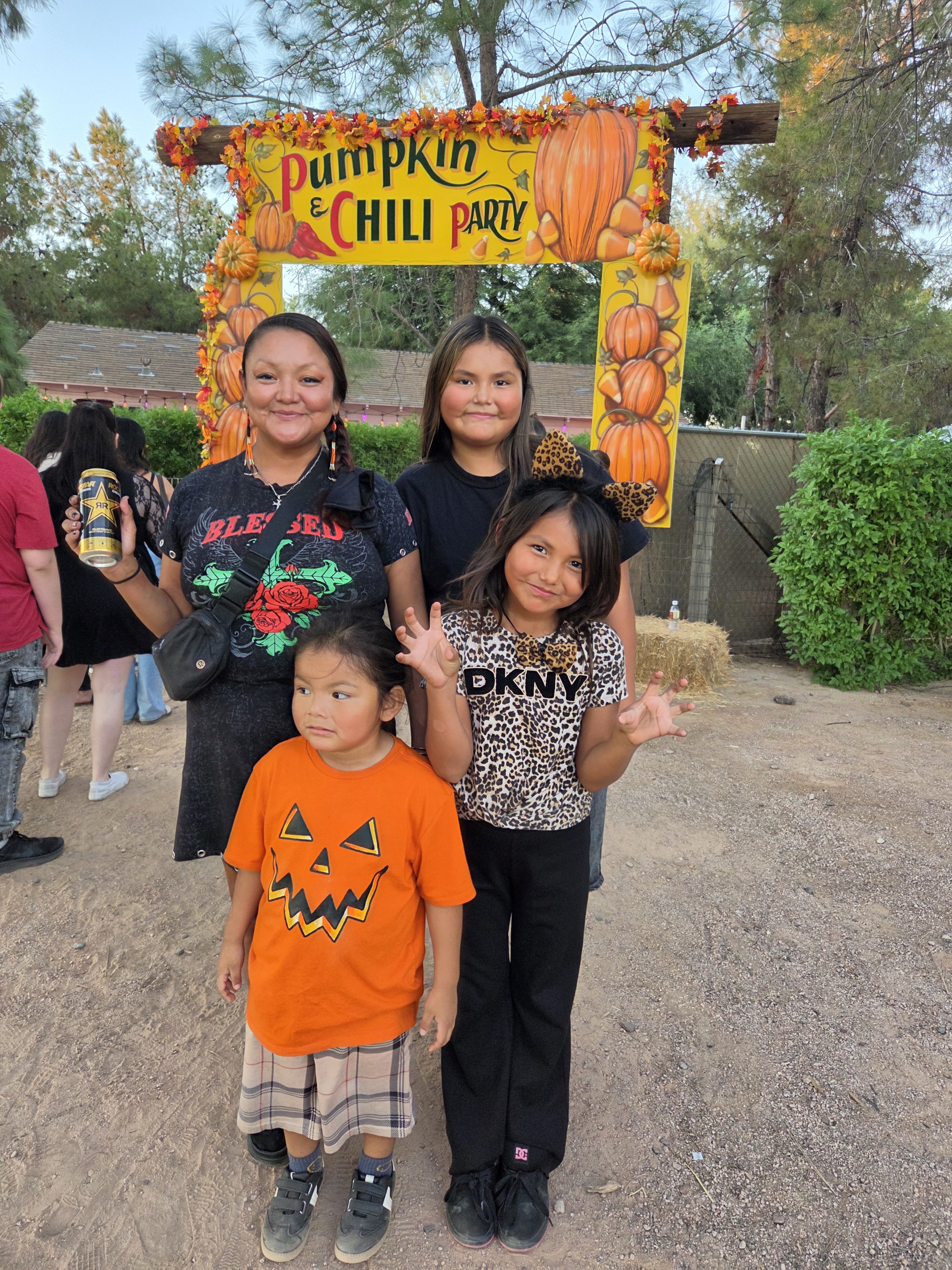 A group of four children and an adult posing in front of a colorful 'Pumpkin & Chili Party' sign decorated with pumpkins and autumn leaves.