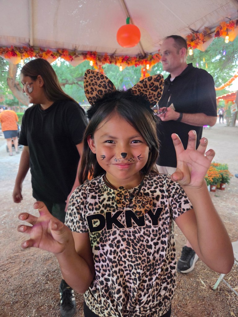 A young girl dressed in a leopard print shirt, wearing leopard ears and face paint, playfully making a claw gesture. In the background, a few other people are seen enjoying the festivities inside a decorated tent.