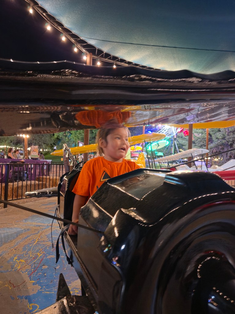 A young child wearing an orange shirt sits in a ride at an amusement park, smiling and enjoying the experience. The ride has black machinery visible, and colorful lights and other rides can be seen in the background.