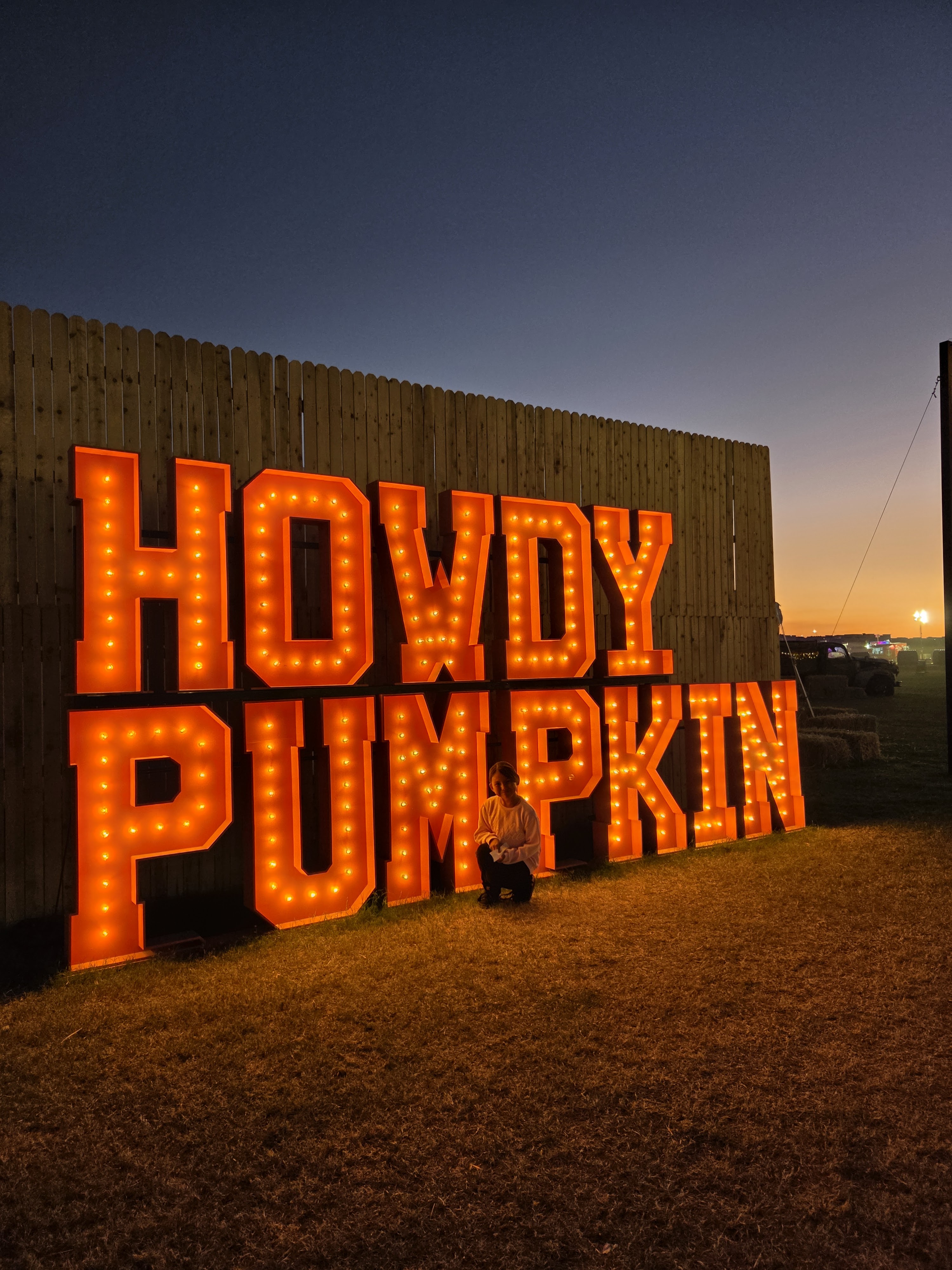 Brightly lit sign displaying the words 'Howdy Pumpkin' against a twilight sky, with a person kneeling beside it.